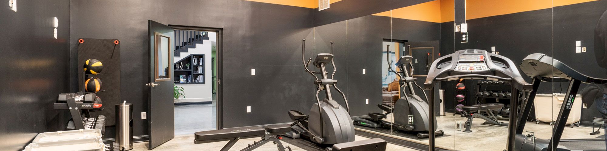 This image shows a small fitness room with equipment including a treadmill, exercise bike, rowing machine, and weights, featuring a mirrored wall.