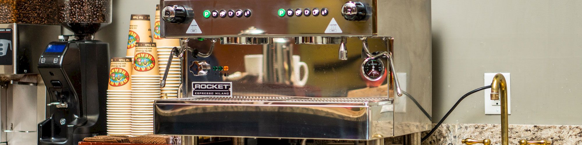 An espresso machine on a counter, surrounded by coffee cups, a grinder, and other coffee-making supplies.