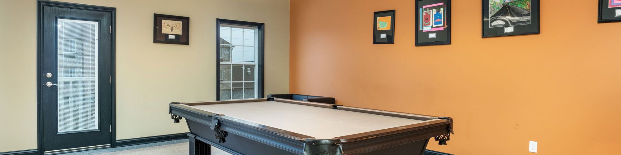 A modern room with a pool table, orange wall, framed artwork, pendant lights, and a glass door. The floor is white and the room is well-lit.