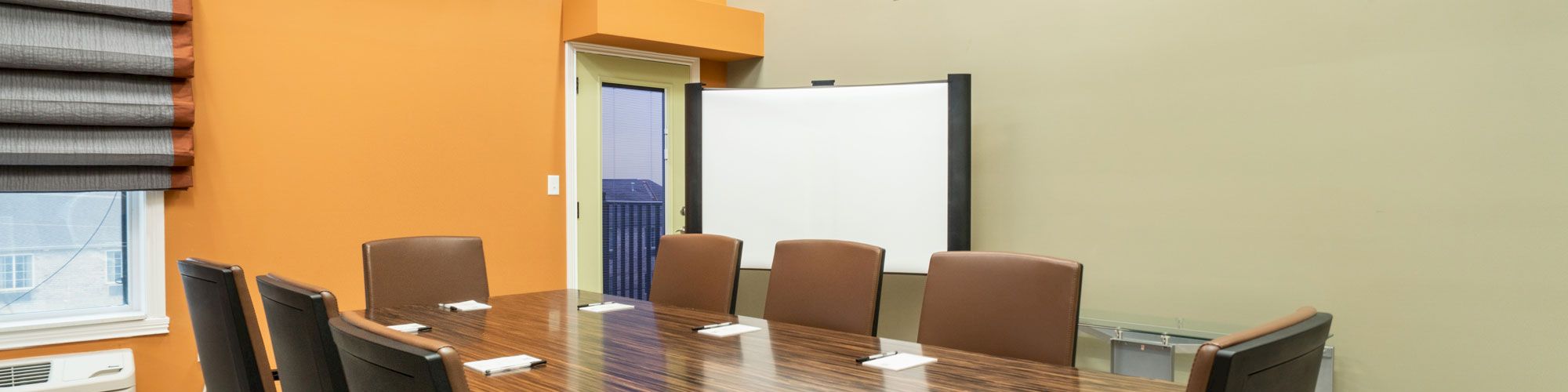 A modern conference room with an orange accent wall, a large wooden table, eight chairs, a whiteboard, and a window with blinds.