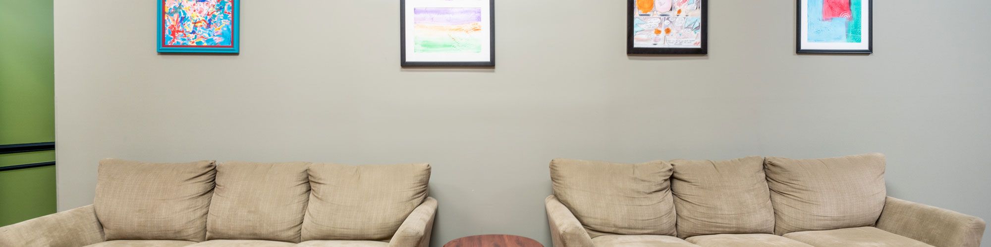 The image shows two beige sofas with matching ottomans, a round wooden table between them, and four framed artworks on the wall behind.