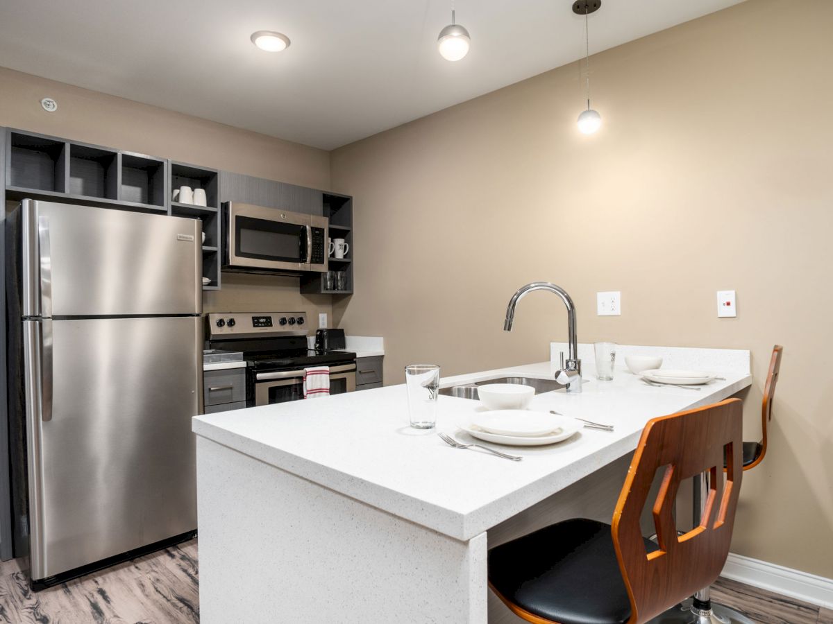 Modern kitchen with stainless steel appliances, a white countertop island with two settings for dining, and wooden flooring ending the sentence.