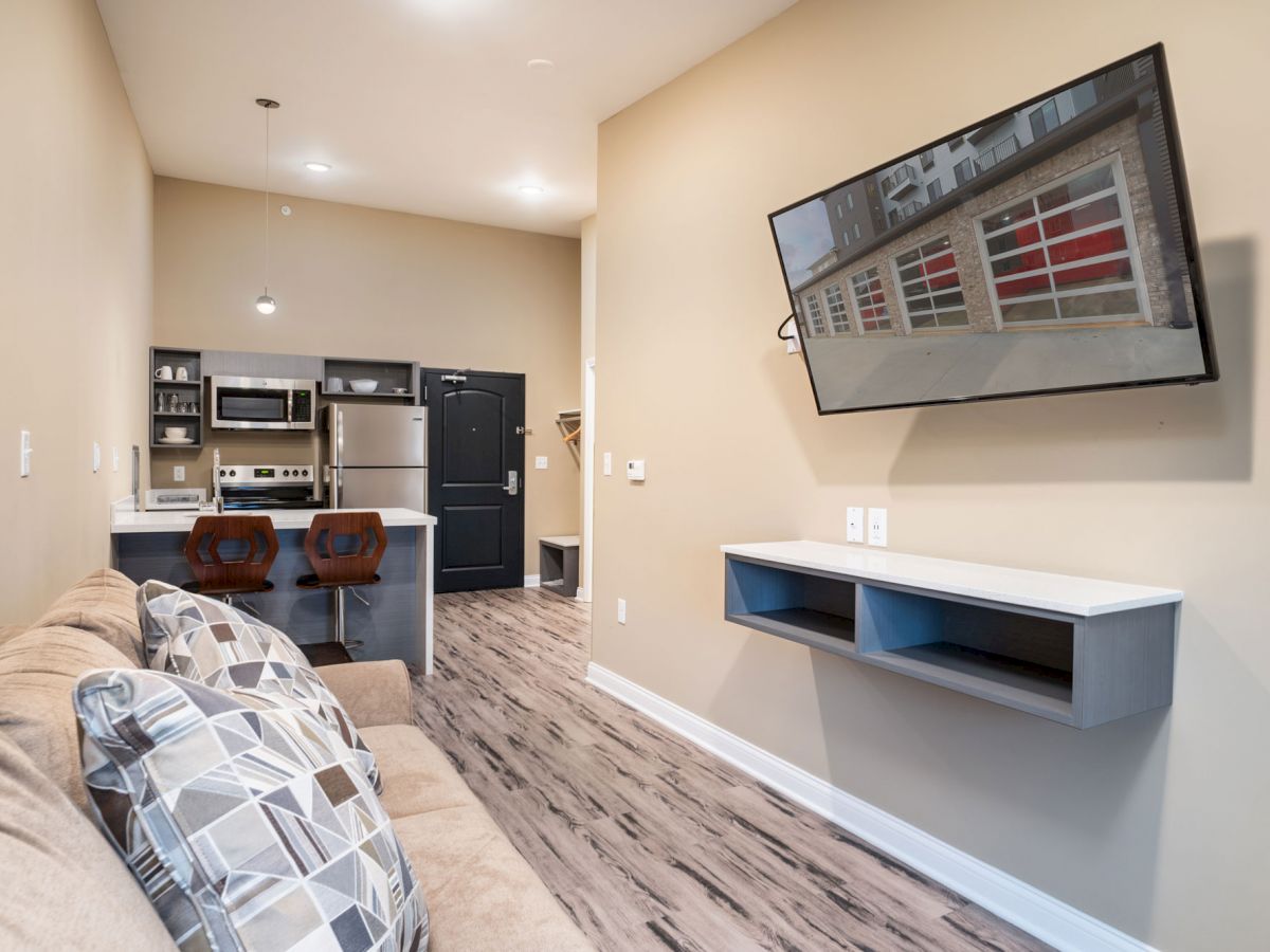 A modern living room with a beige couch, patterned pillows, a wall-mounted TV, a small kitchen area, and two bar stools by the counter.