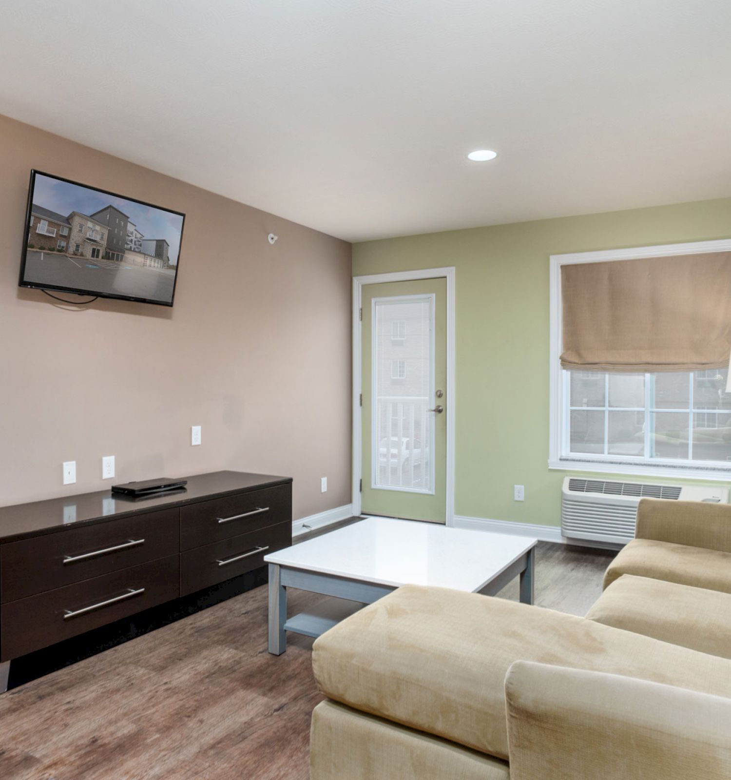 A living room with a beige couch, coffee table, TV on the wall, console, window, and a lamp near a door leading outside.