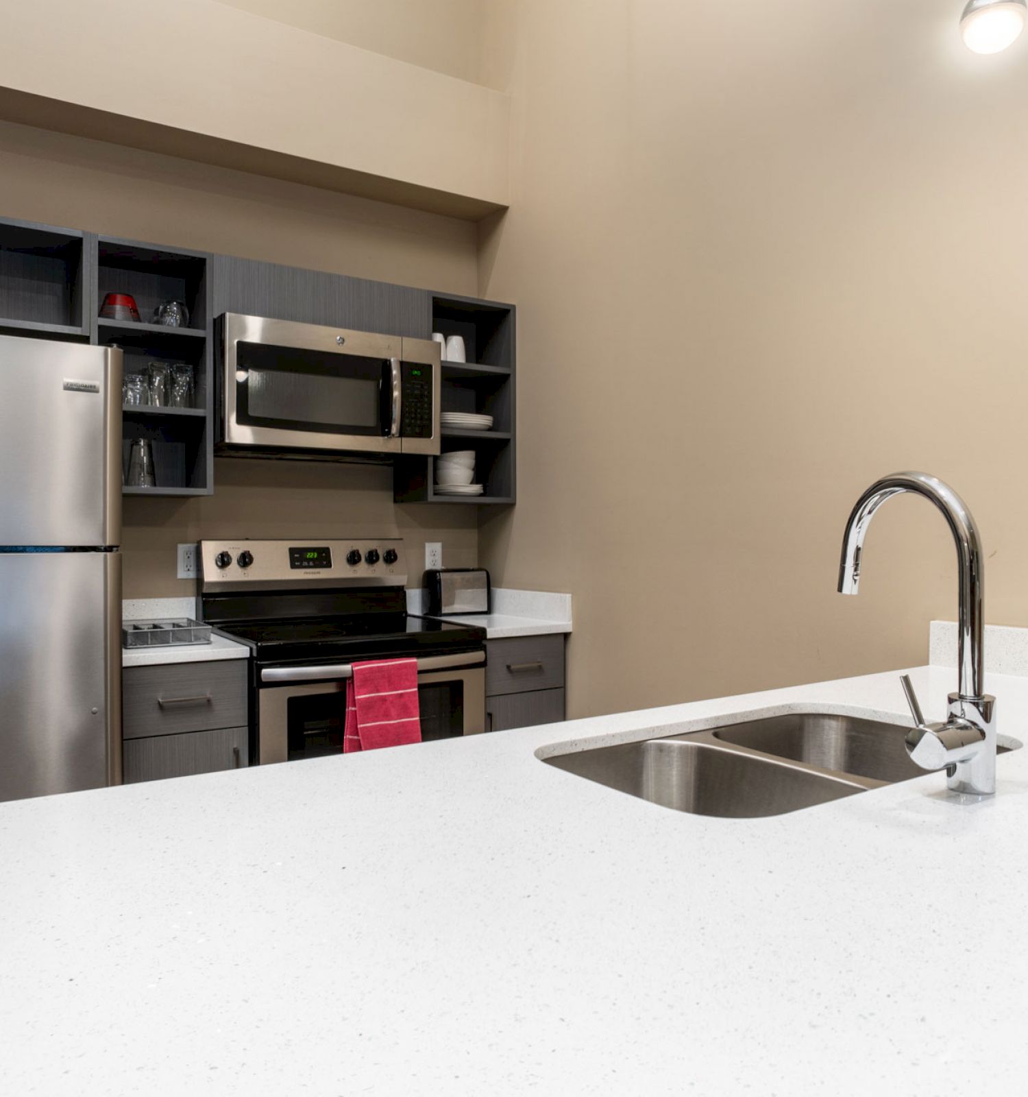 A modern kitchen with stainless steel appliances, white countertops, a double sink, and a wall-mounted microwave. A red towel hangs on the oven.