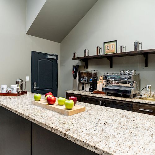 A coffee station with a countertop featuring apples, a coffee machine, mugs, and shelves with containers above.