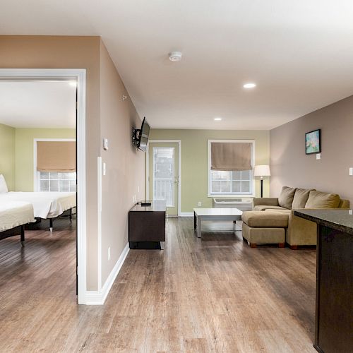 A modern living space with wood flooring, a kitchen counter, a comfortable sofa, and a bedroom visible through an open door.