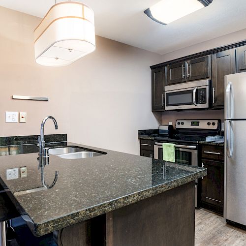 A modern kitchen with dark cabinets, stainless steel appliances, an island with a sink, two stools, and pendant lighting.