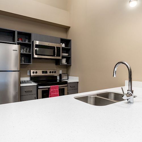 A modern kitchen with stainless steel appliances, a double sink, a microwave, and an oven. The countertop is white, and the cabinets are gray.