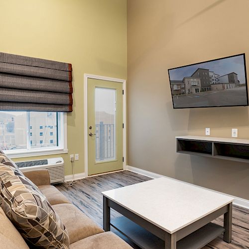 A furnished room with a sofa, coffee table, wall-mounted TV, door, window with blinds, and wooden floor.