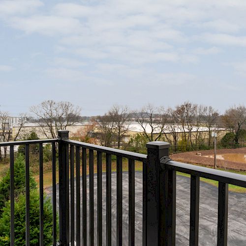 The image shows a view from a balcony with a black railing, overlooking an area with trees, a field, and some buildings in the distance.