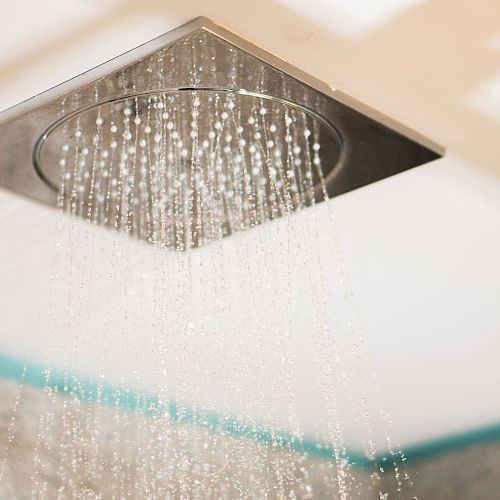 A ceiling-mounted square rain shower head with water spraying down in a bathroom.