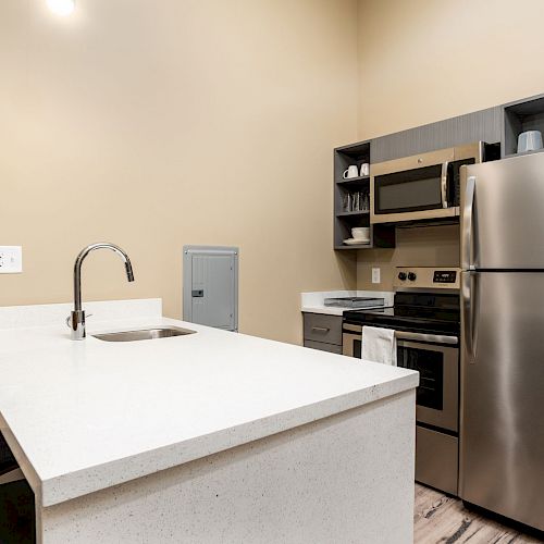 A modern kitchen with a countertop, sink, stool, stainless steel refrigerator, microwave, and oven, set against a beige wall, with shelving.