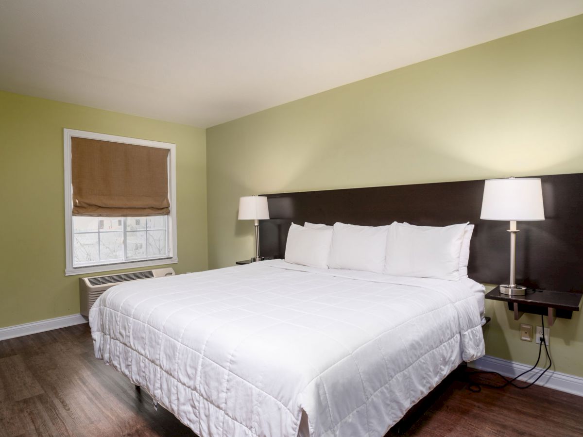 A bedroom with a large bed, two bedside tables with lamps, a window with a closed shade, green walls, and wooden flooring.