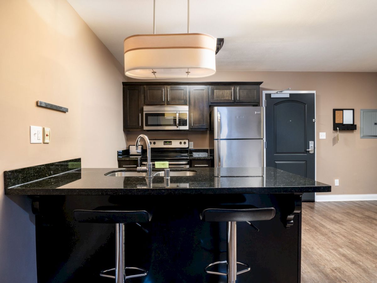 A modern kitchen with a black countertop island, two stools, stainless steel appliances, dark cabinetry, and a minimalist design.