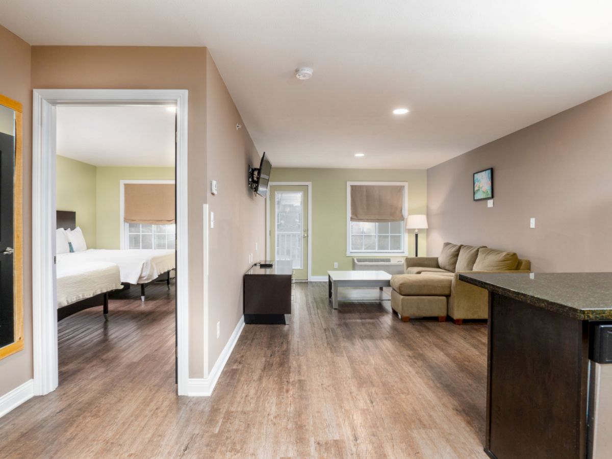 This image shows a modern living room with a sectional sofa, a TV on the wall, wooden floors, and an adjacent bedroom with a bed visible.