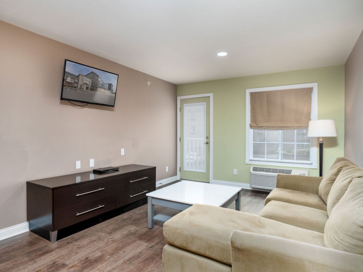 A cozy living room with a beige couch, wall-mounted TV, coffee table, and a window with a shade, ending the sentence.