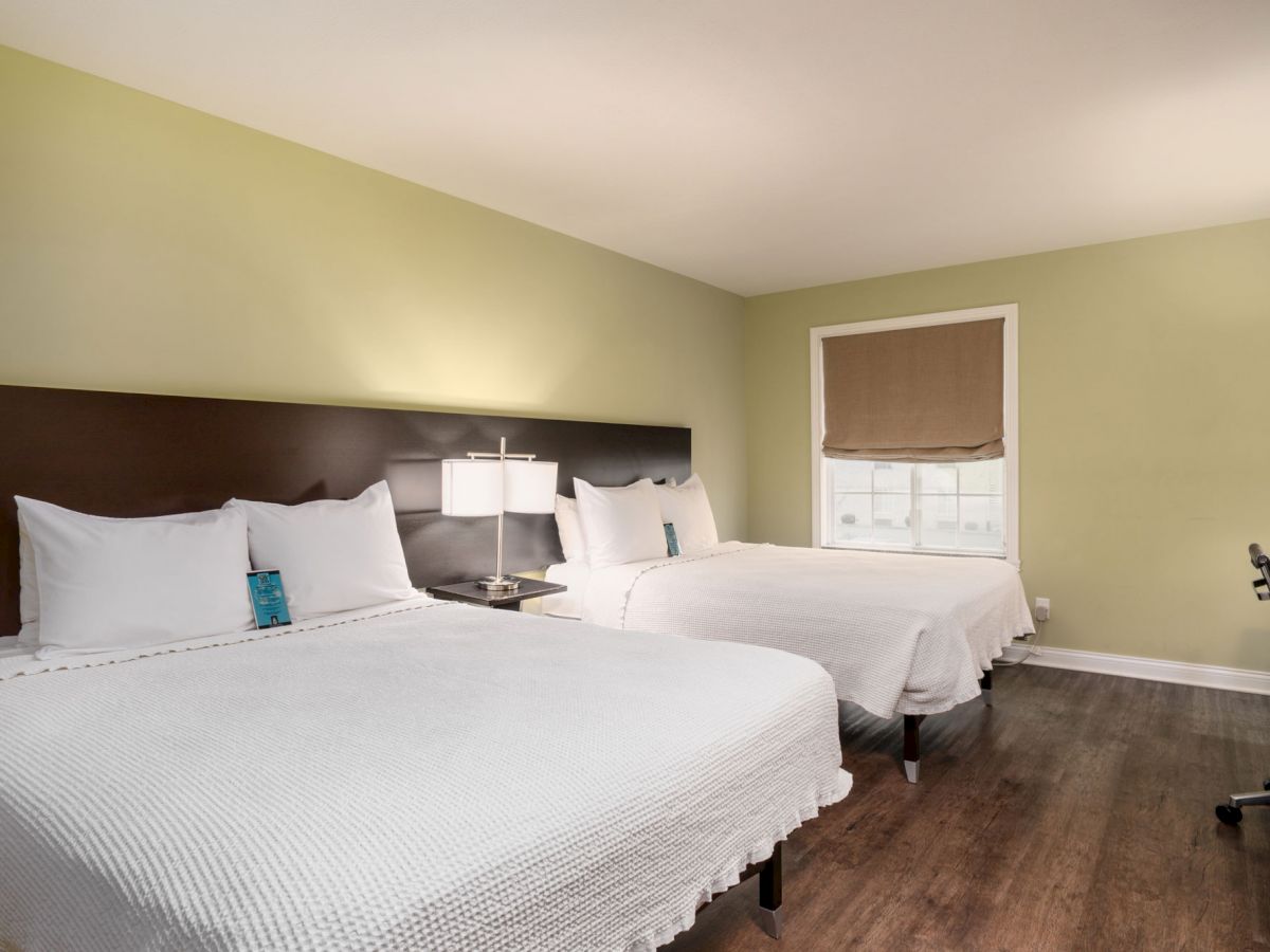 The image shows a hotel room with two double beds, white bedding, a small desk, and a window with a blind, on a wooden floor.