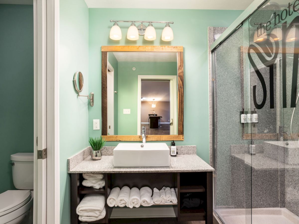 A modern bathroom features a sink, mirror, toilet, towels, and a glass-walled shower with a visible logo. A doorway opens to another room.