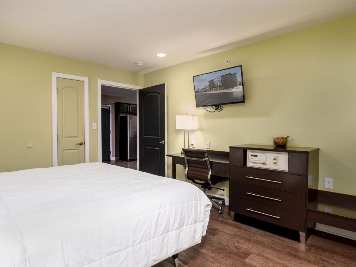 A neatly arranged bedroom with a white bedspread, a desk with a chair and lamp, a wall-mounted TV, and a dark dresser against a light green wall.