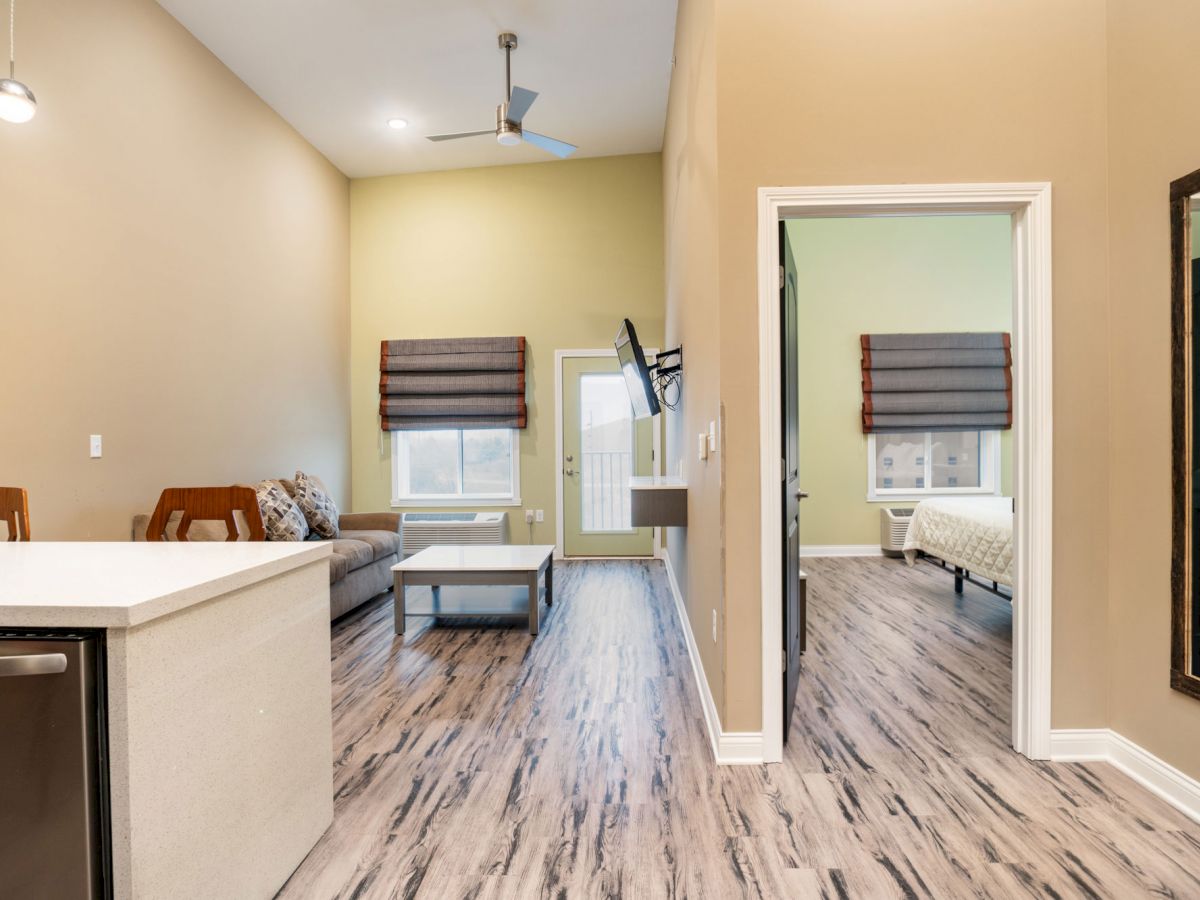 This image shows a modern interior with a living area, open kitchen, and a bedroom. There's a blend of wood flooring, neutral colors, and minimalist decor.