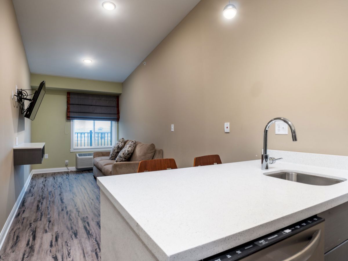 The image shows a modern, narrow living space with beige walls, a mounted TV, a sofa, and a kitchen area. A large window brings in natural light.