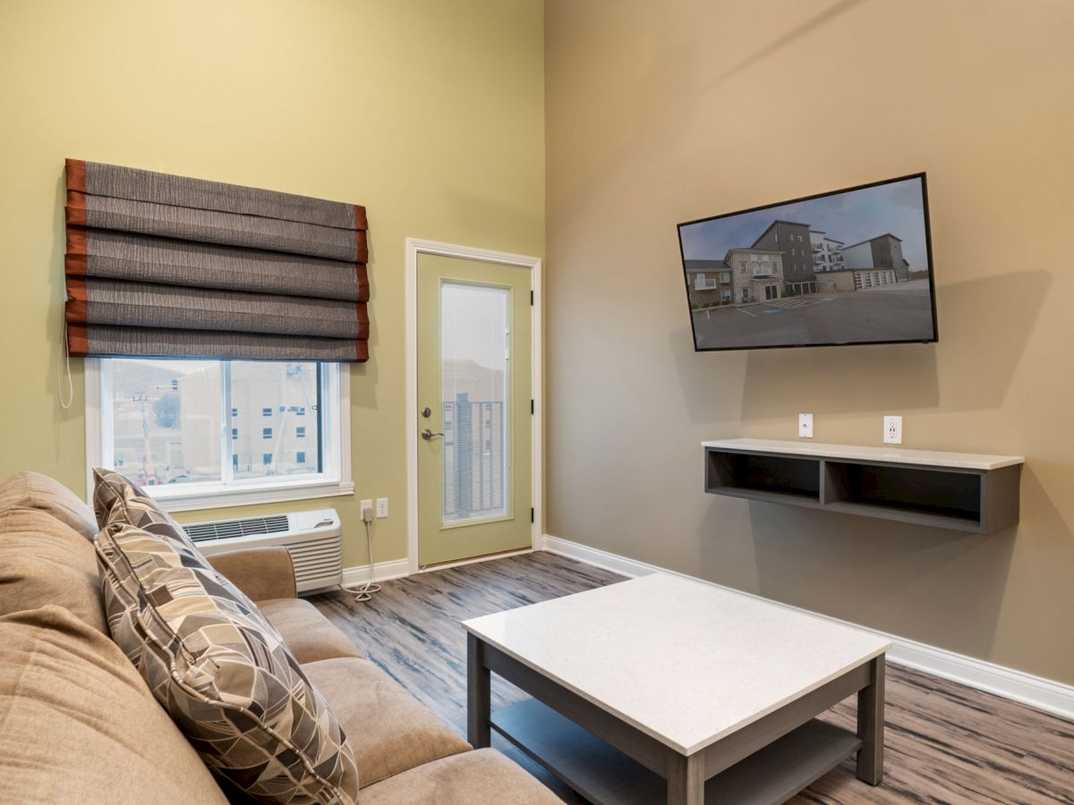 A cozy living room with a couch, coffee table, mounted TV, window with blinds, door, and a small storage shelf beneath the TV ending the sentence.