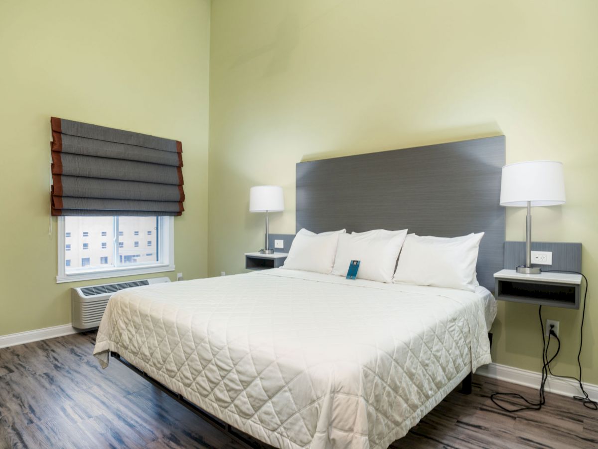 A modern bedroom with a large bed, two side tables with lamps, wooden flooring, a window with blinds, and light green walls.