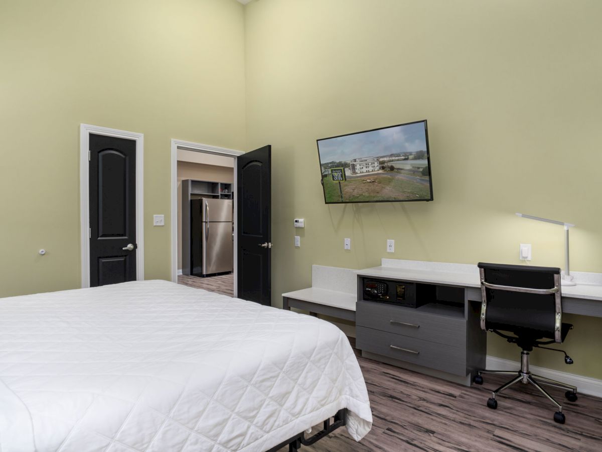 A bedroom with a white quilted bed, desk area with a chair and lamp, wall-mounted TV, two doors, one showing an adjacent stainless steel appliance.