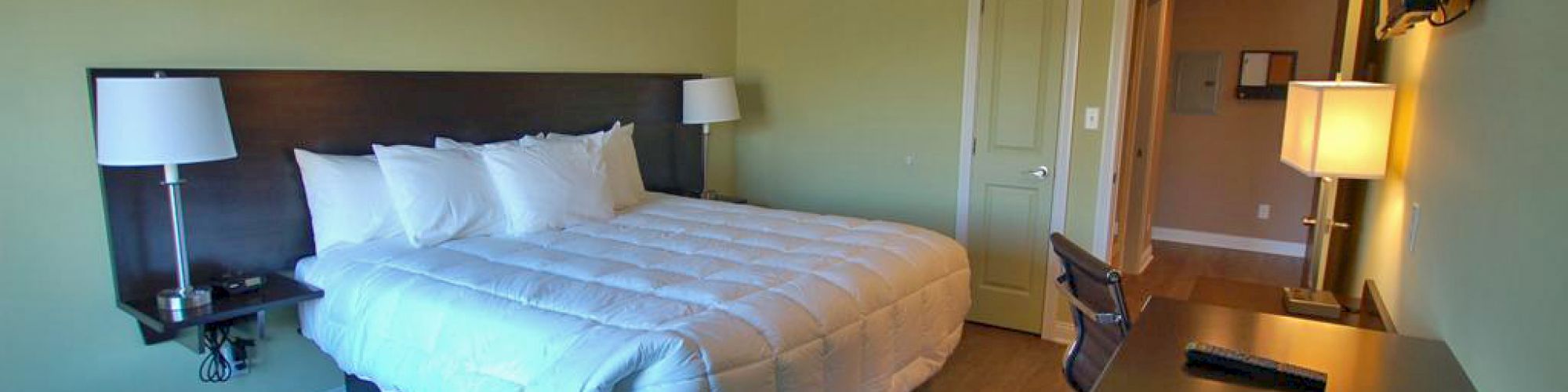 A modern hotel room with a bed, two bedside lamps, a desk, chair, and wall-mounted TV. The room has light walls and a wood floor.