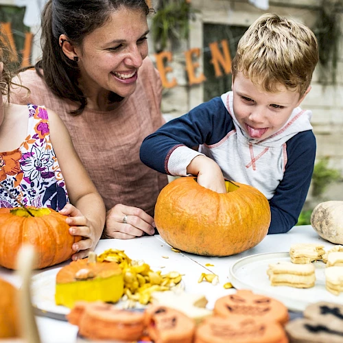A woman and two children are carving pumpkins together at a table filled with more pumpkins and Halloween-themed treats, enjoying themselves.