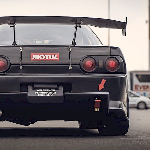 The image shows the rear view of a modified black car with a Motul sticker, red arrow, and a tow hook on its bumper, parked in an open area.