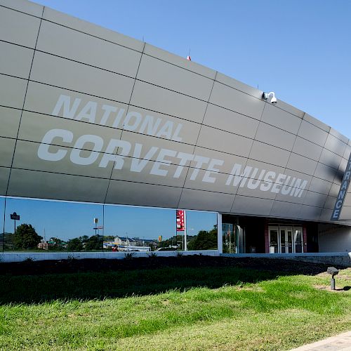 The image shows the exterior of the National Corvette Museum, a modern building with angled walls and large glass windows reflecting the surroundings.