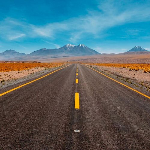 A straight road stretches into the distance, flanked by a dry desert landscape with mountains under a bright blue sky in the background.