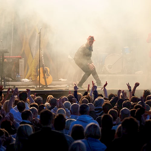 A band is performing on stage with a keyboard, drums, and guitar, surrounded by smoke, while a large crowd enjoys the music.