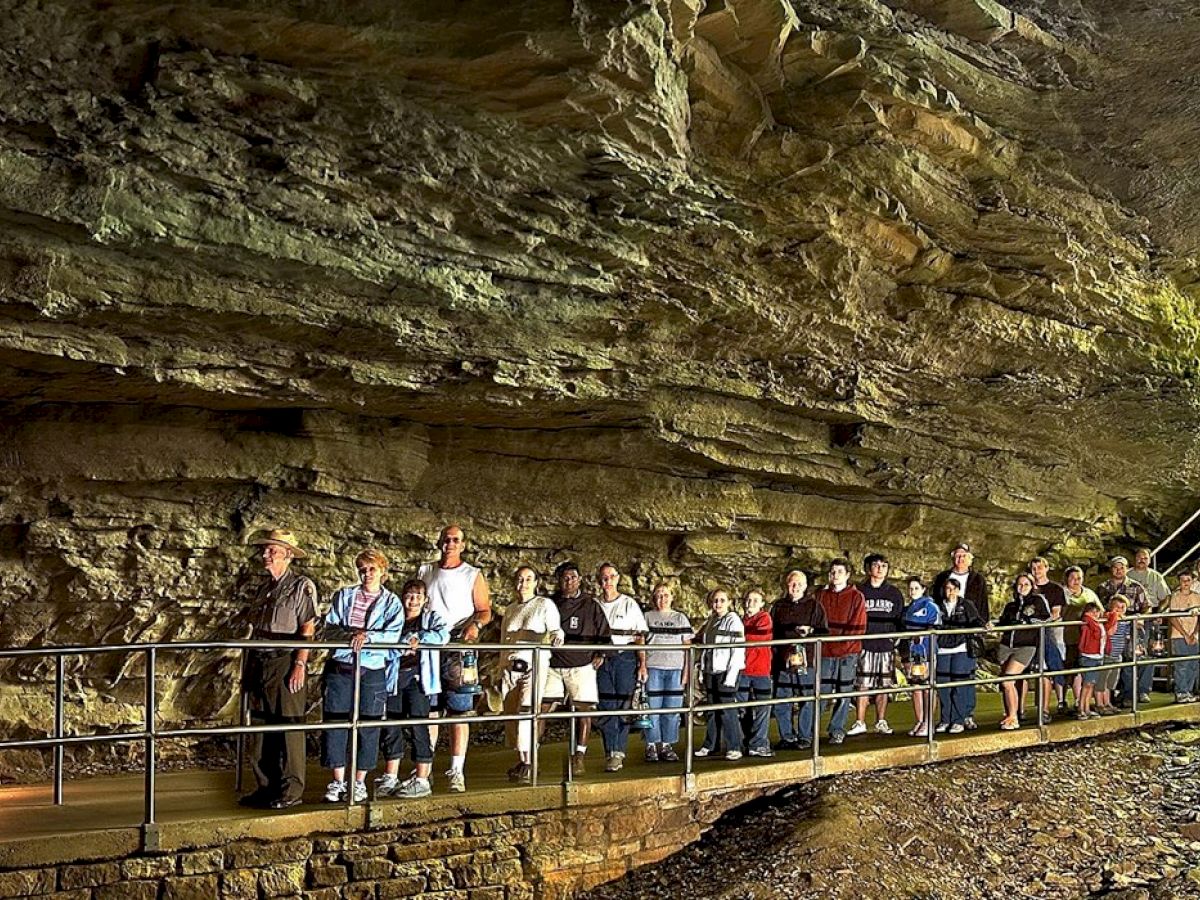 A group of people walk through a cave on a raised walkway, with sunlight streaming in through the large entrance opening.