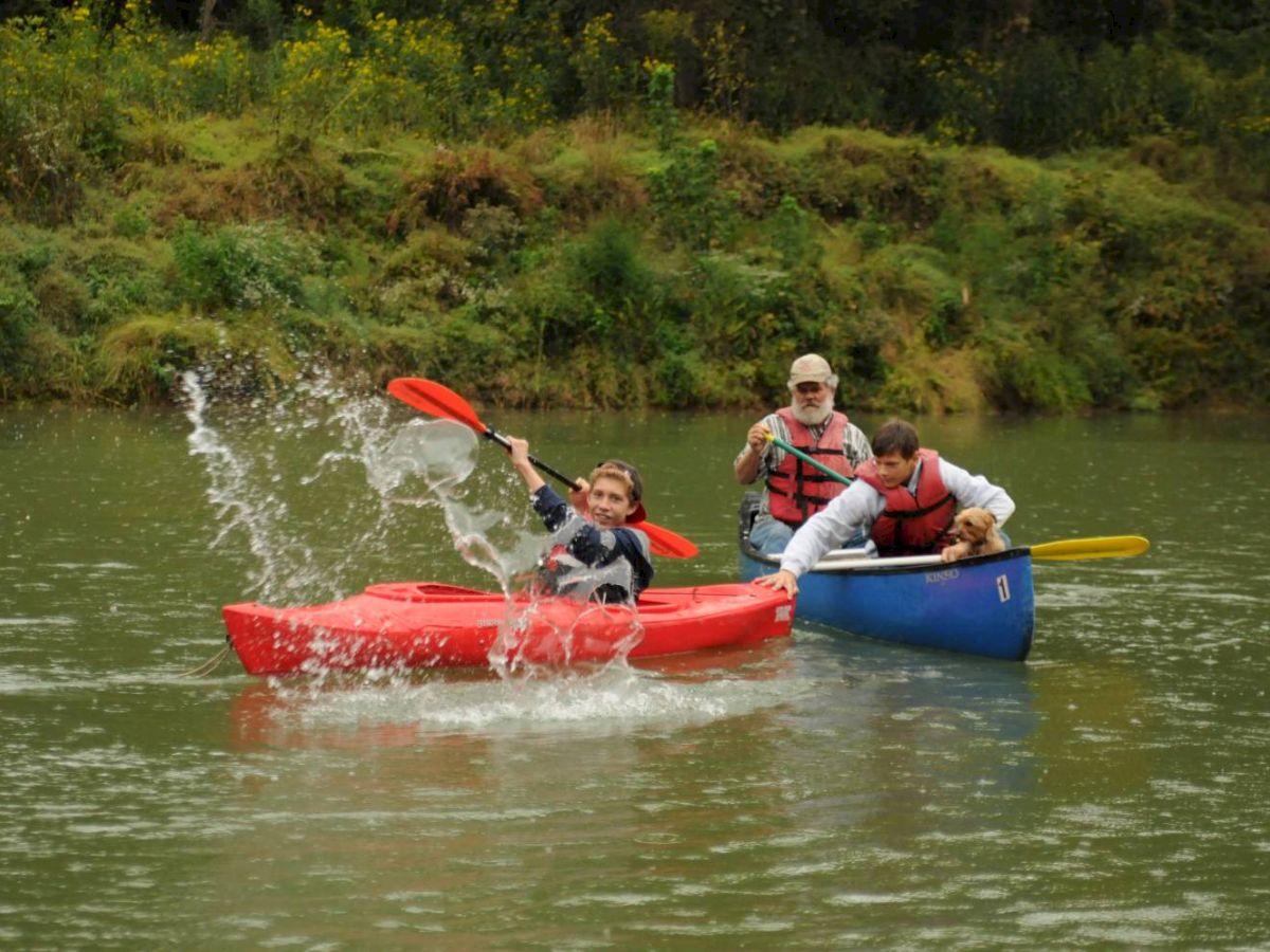 Three people are kayaking in a river, with one person splashing water using their paddle. They are all wearing life jackets.