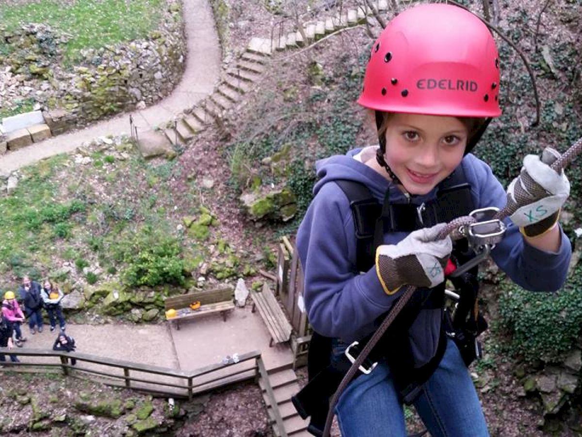 A person is mid-climb on a rope course, wearing a red helmet and safety gear. Others watch from below on a pathway surrounded by greenery.