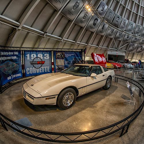 The image shows a white car displayed in a museum, surrounded by a circular barrier, with informational banners and other cars in the background.