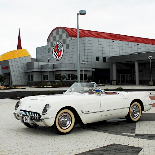 A white classic car is parked in front of a modern building with a yellow and red roof, and a checkered shield logo is visible on the building.