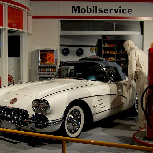 A classic car at a vintage gas station exhibit, with a mannequin attendant and retro fuel pumps in front of the Mobilservice station.