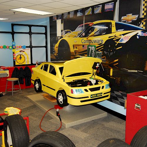 A kids' playroom with a mini yellow car on a stand, toy tools, tires, racing-themed decor, and a large wall mural of race cars.