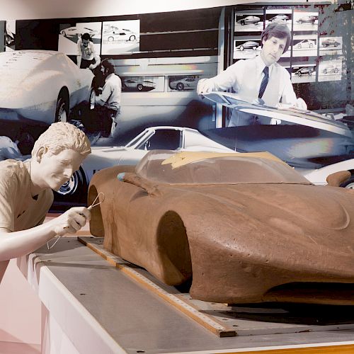 A statue of a person is sculpting a car model in the foreground, with a backdrop showing people working on car designs.