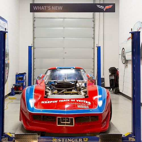 A red racing car is on a lift in a garage, surrounded by automotive equipment and decor. The car hood is open, showing the engine.