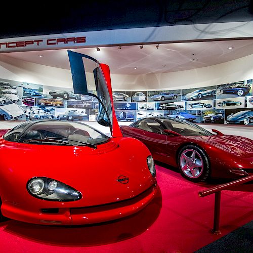 The image shows a car showroom featuring two red concept cars on display, with a background of various car designs and the sign 