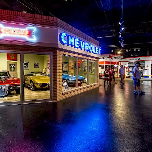 An indoor exhibit featuring classic cars and a neon Chevrolet sign, with visitors walking around the area.