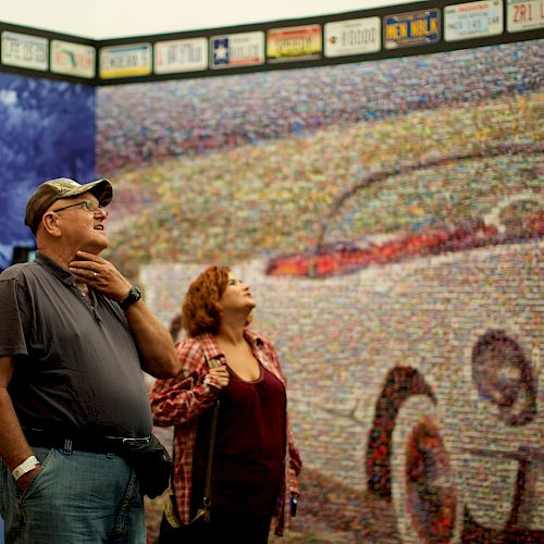 Two people are admiring a large mosaic depicting a car, with various license plates displayed above and an informational display titled 