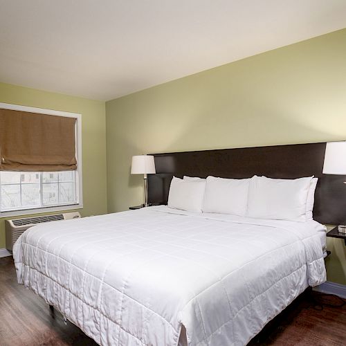 A bedroom with a large bed, two bedside lamps, a window with a brown shade, and light green walls. The floor is dark wood.