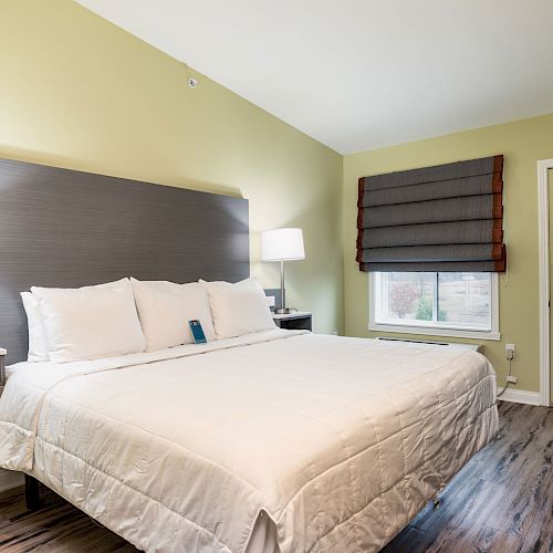 A modern hotel room with a large bed, two bedside tables with lamps, a window with a view, and a door to the outside. The room has wooden floors.