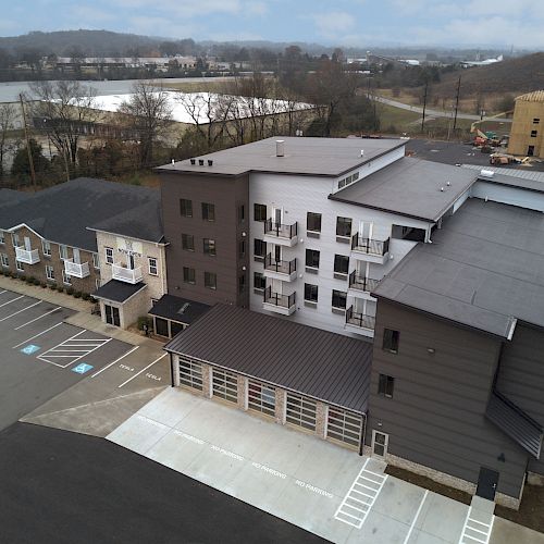 Aerial view of a modern apartment complex with parking spaces, adjacent buildings, and surrounding landscape including trees and a body of water visible.